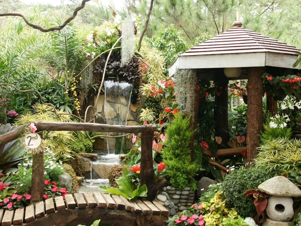 A lush garden scene featuring a small waterfall, a wooden bridge, and a gazebo surrounded by abundant greenery and flowers.