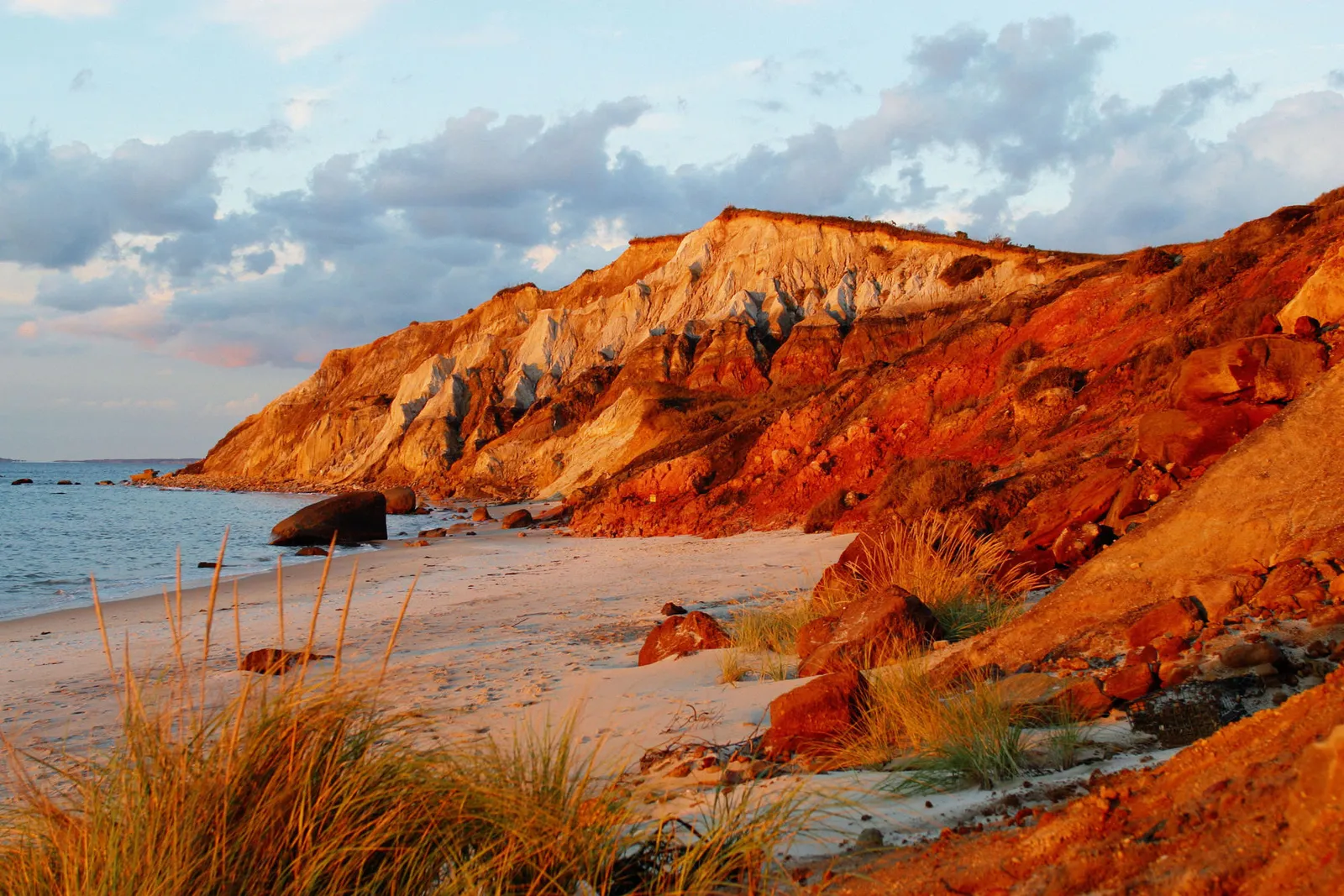 A sandy beach leads to colorful cliffs bathed in golden sunset light, with the ocean and clouds in the background.