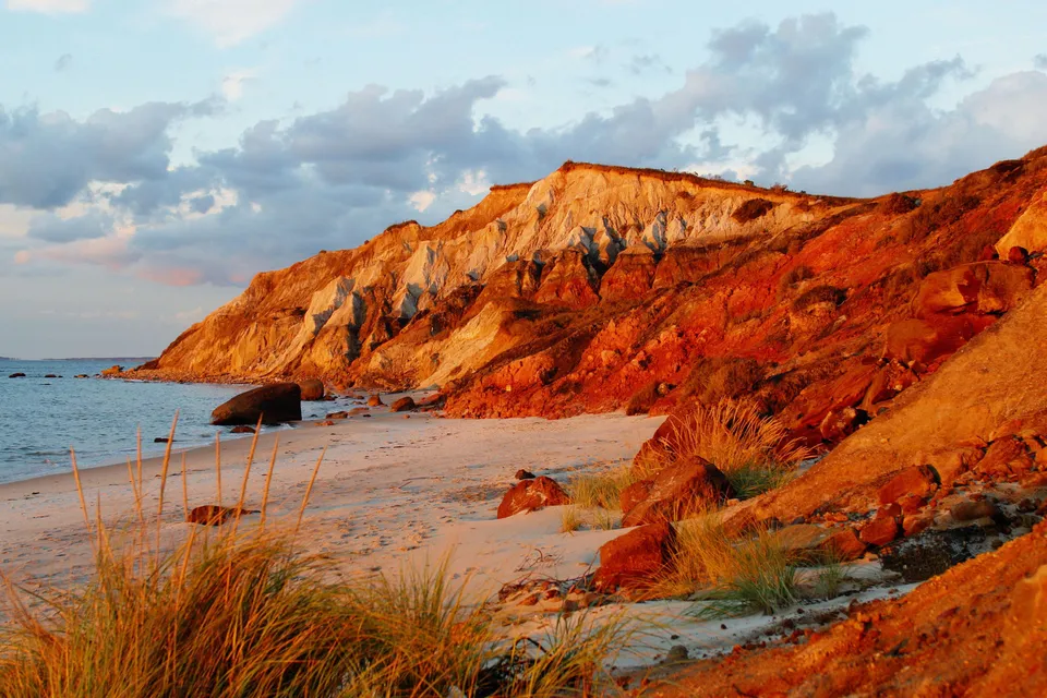 A sandy beach leads to colorful cliffs bathed in golden sunset light, with the ocean and clouds in the background.