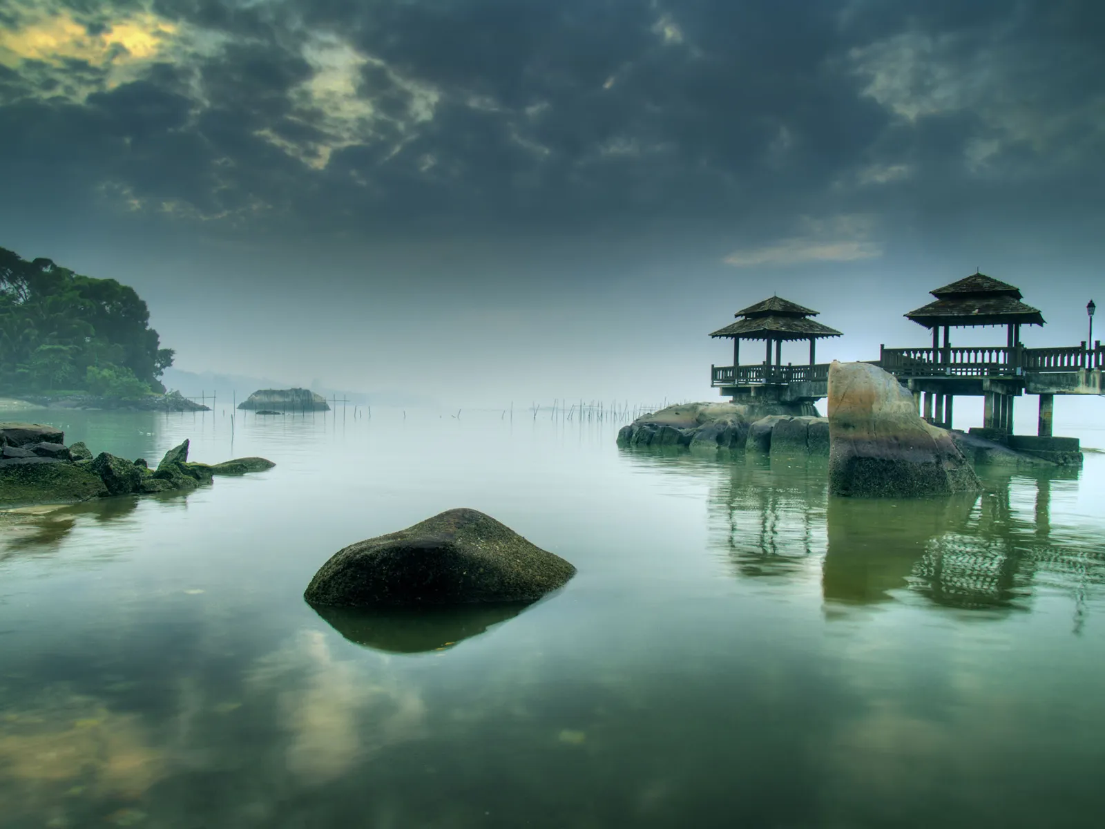 Two wooden gazebos stand on stilts over calm water, with rocks and trees in the background under a cloudy sky.