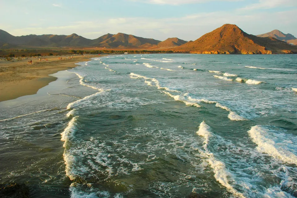 Gentle waves roll onto a sandy beach with mountains in the background during sunset.