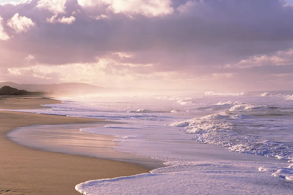 A serene beach scene with gentle waves washing onto the sand under a cloudy sky.