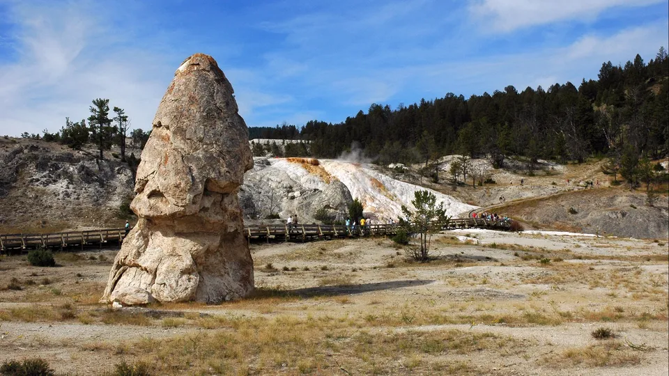 A large, weathered rock formation stands in the foreground with a steaming geothermal terrace and boardwalk in the background.