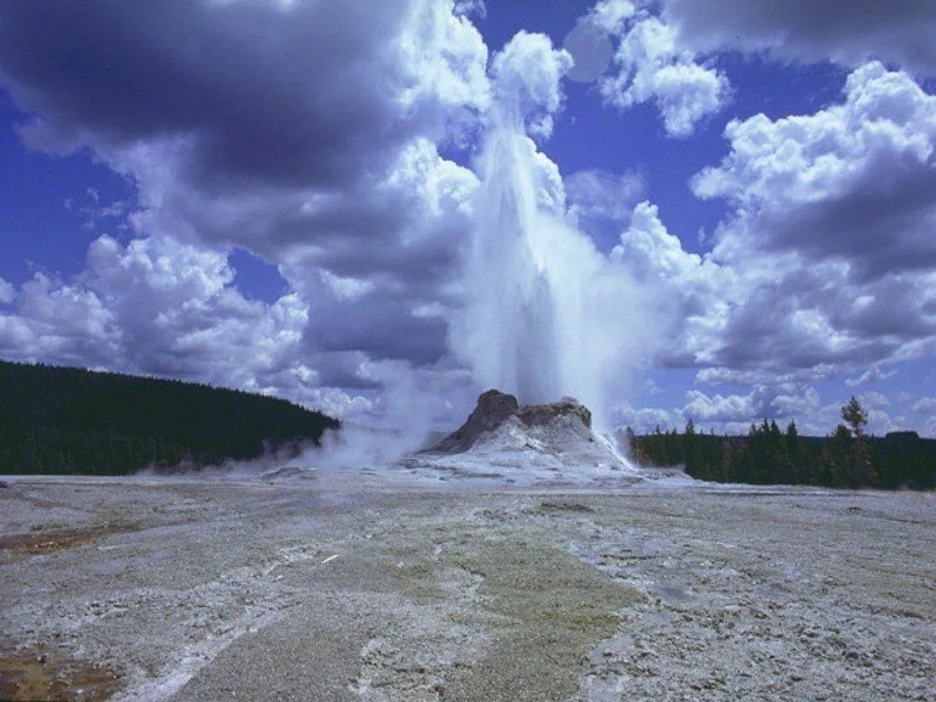 A geyser erupts with a tall plume of water and steam against a backdrop of dramatic clouds and blue sky.