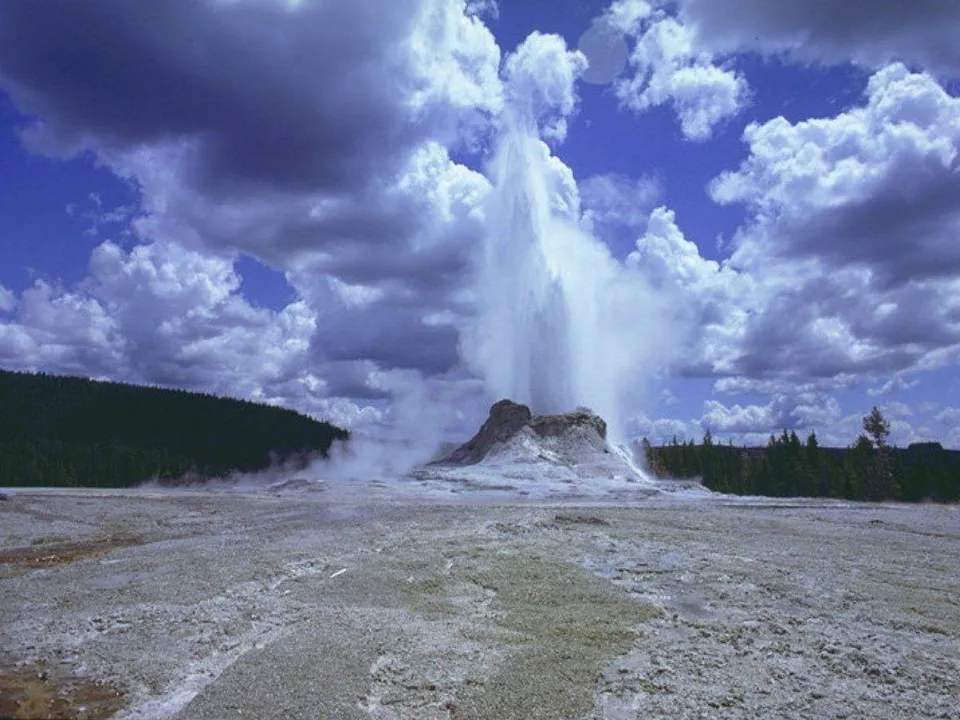 A geyser erupts with a tall plume of water and steam against a backdrop of dramatic clouds and blue sky.
