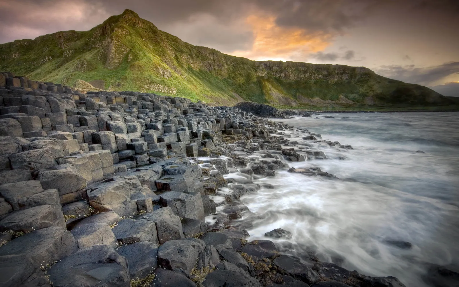 Hexagonal basalt columns line the coast with a green hill and dramatic sky in the background.