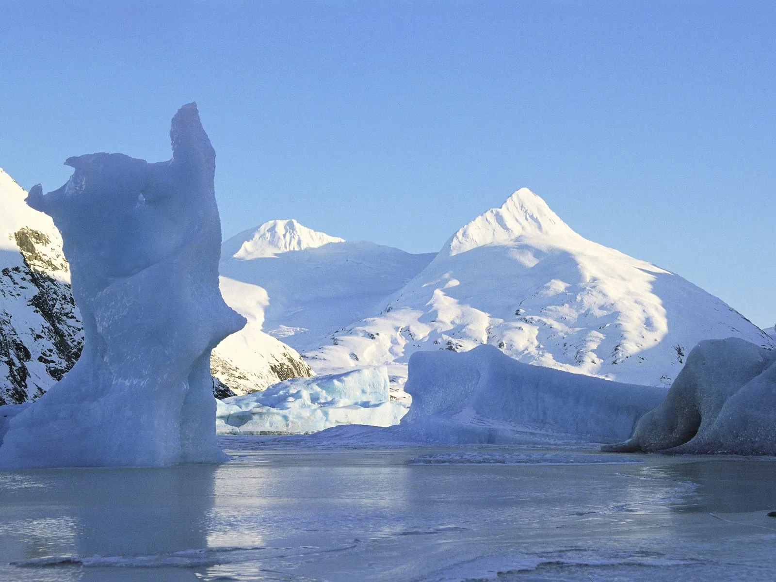 A striking ice formation stands in a frozen body of water, with snow-covered mountains in the background under a clear blue sky.