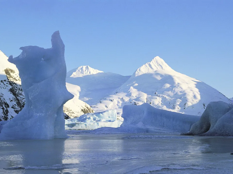 A striking ice formation stands in a frozen body of water, with snow-covered mountains in the background under a clear blue sky.