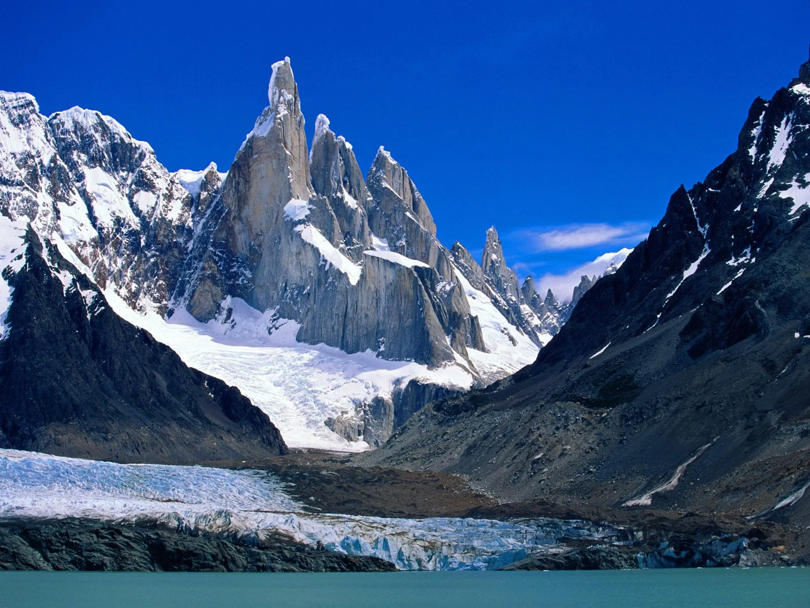Jagged, snow-capped mountains rise above a large glacier and a calm blue lake under a clear sky.