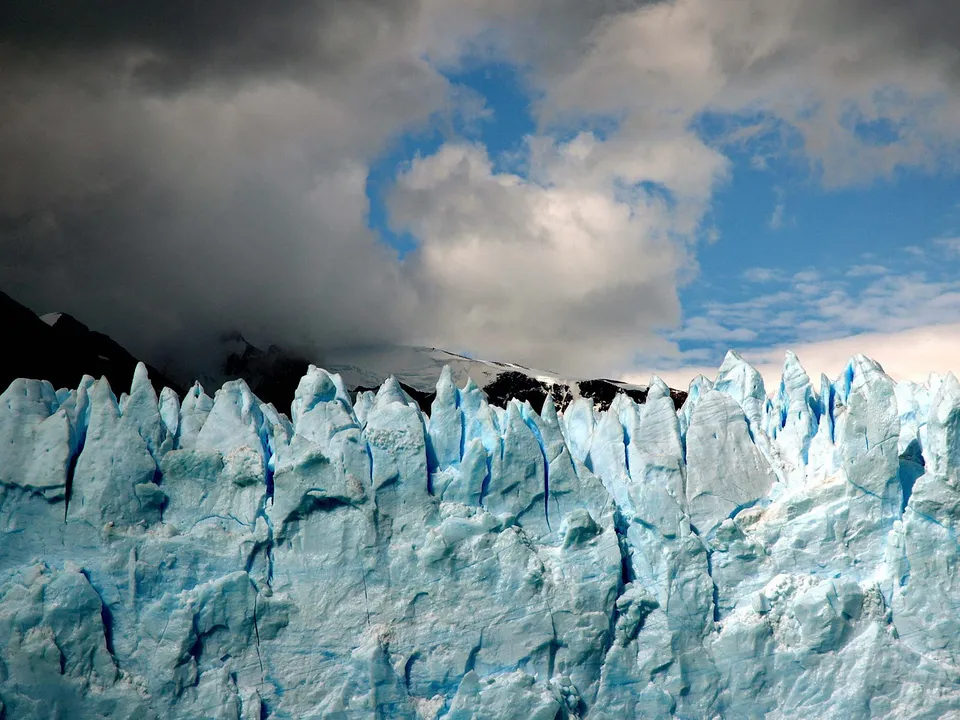 Close-up of a bright blue glacier face with jagged ice formations against a dramatic sky with dark clouds and patches of blue.