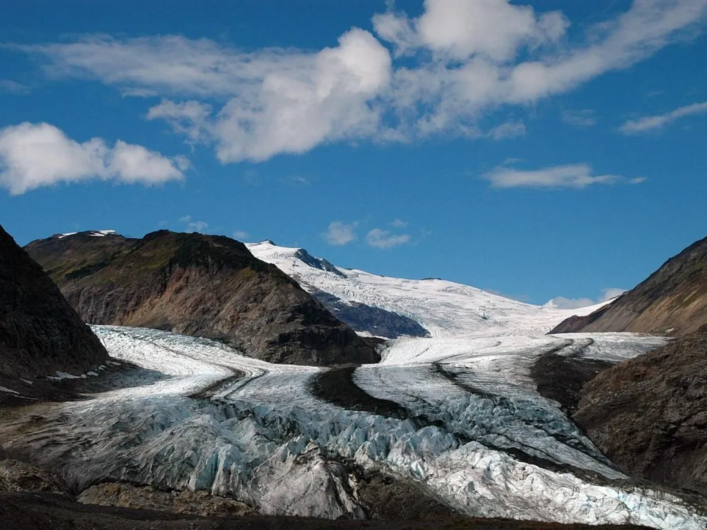 A vast glacier carves its way through a rocky mountain valley under a blue sky with scattered clouds.