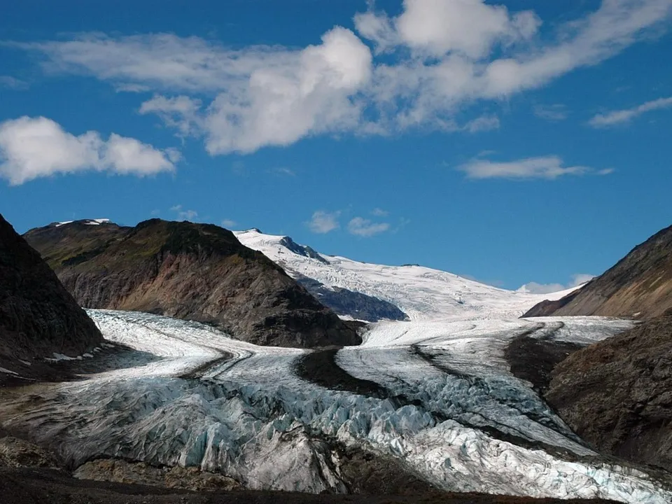 A vast glacier carves its way through a rocky mountain valley under a blue sky with scattered clouds.