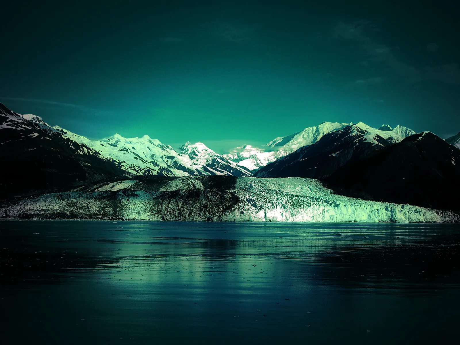 A vast glacier flows down from snow-capped mountains towards a dark, reflective body of water under a teal sky.