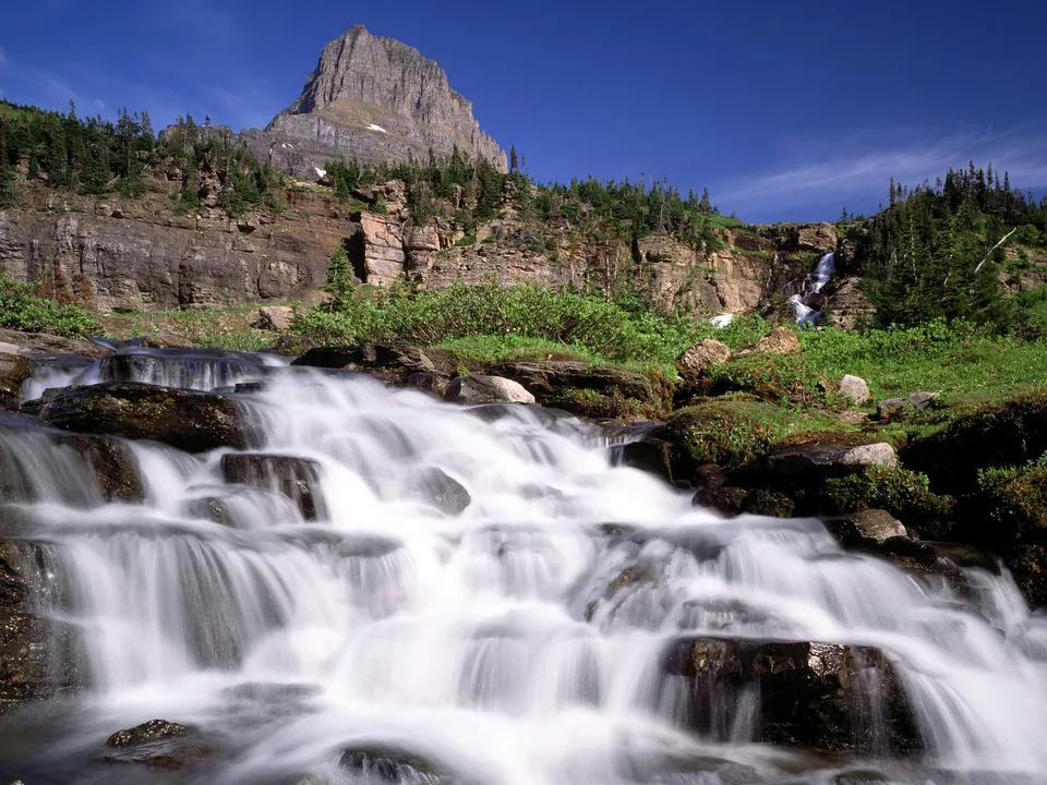A wide waterfall cascades down rocks in Glacier National Park under a bright blue sky.