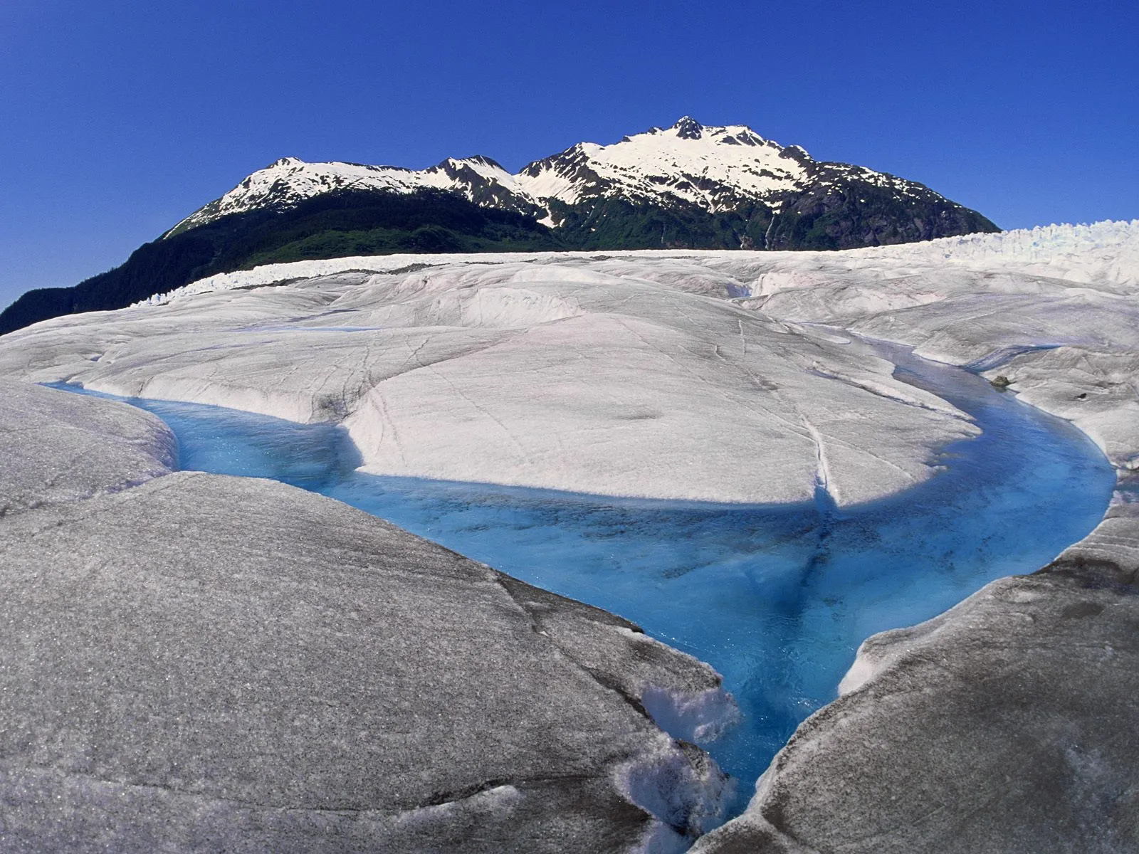 A bright blue river carves a path through a vast, textured glacier, with snow-capped mountains in the background under a clear blue sky.
