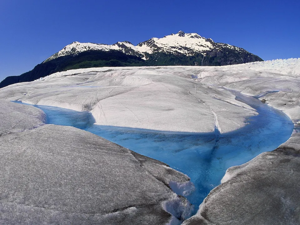 A bright blue river carves a path through a vast, textured glacier, with snow-capped mountains in the background under a clear blue sky.