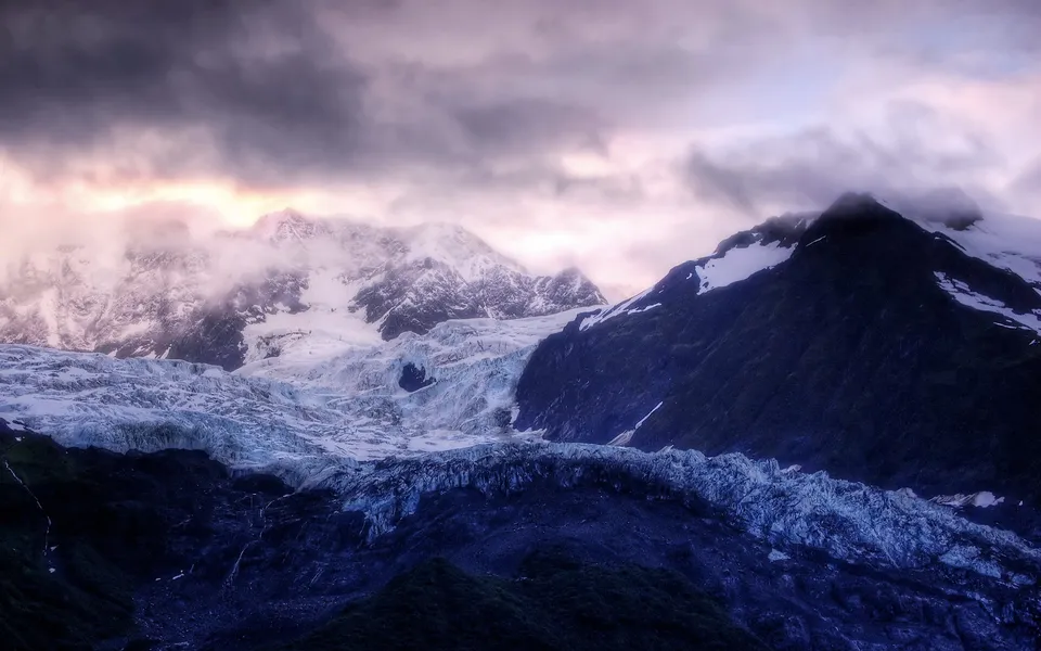 A vast glacier flows down a mountain valley under a dramatic, cloudy sky with hints of pink and yellow light.