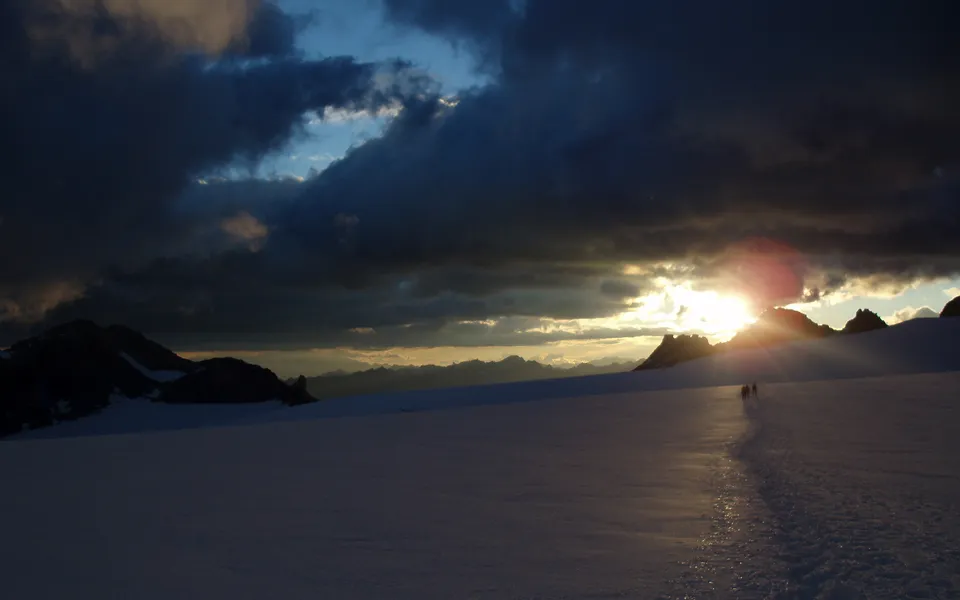 Silhouetted hikers walk across a vast, snow-covered glacier towards a dramatic sunset breaking through dark clouds.