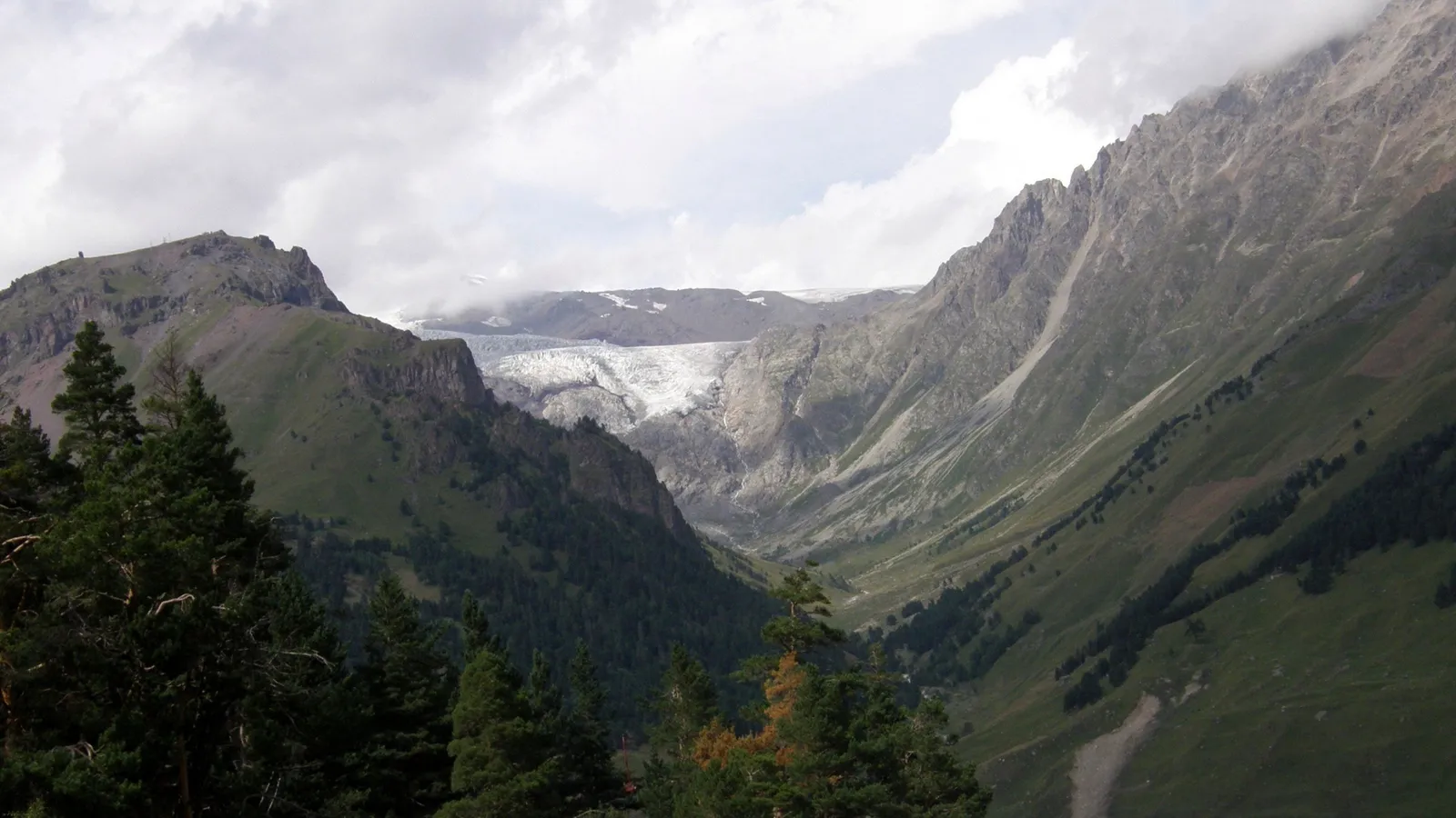 A wide valley with a glacier at the far end, flanked by steep, green mountains under a cloudy sky.