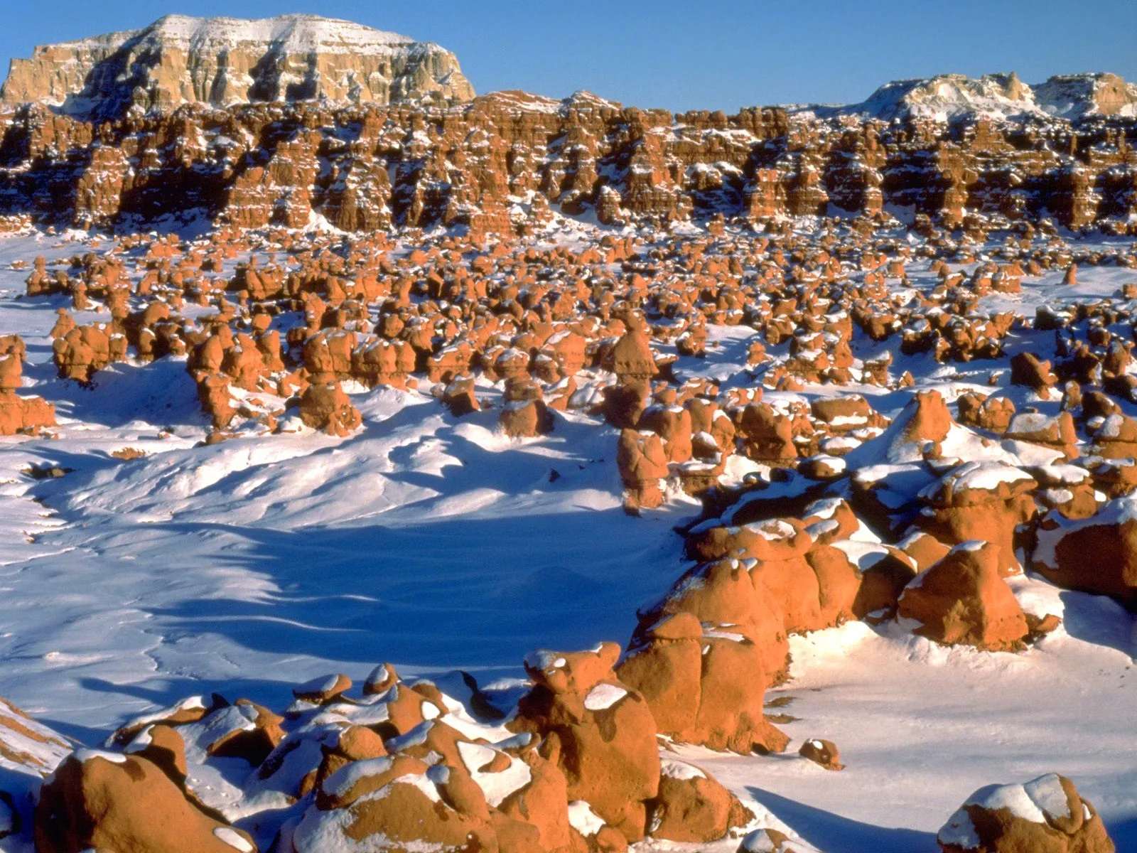 A vast landscape of orange hoodoos and rock formations dusted with snow under a clear blue sky.