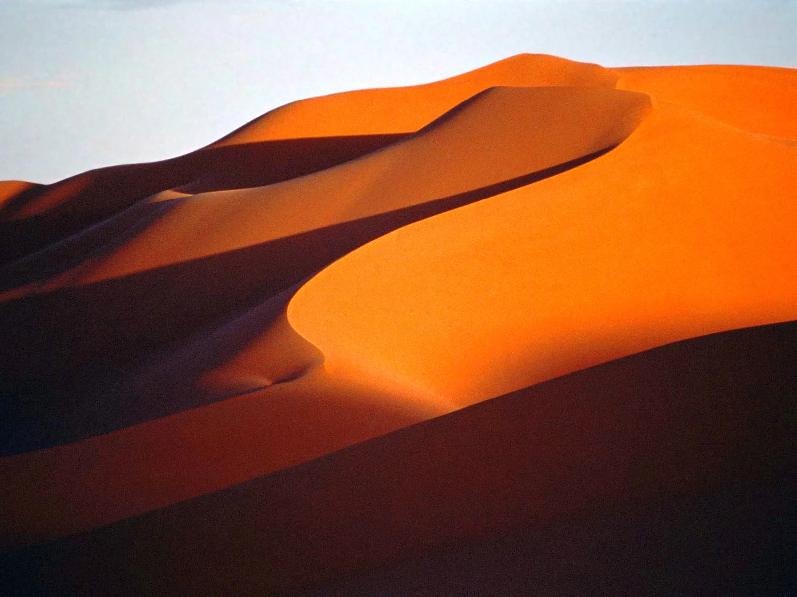 Curving sand dunes in warm orange and deep shadow tones, with a pale sky above.