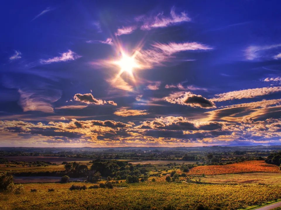 A vast rural landscape with golden fields and trees under a vibrant blue sky with dramatic clouds and a bright sun.