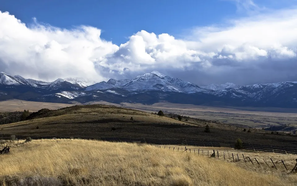 A vast landscape with golden dry grass in the foreground, rolling hills, and a snow-capped mountain range under a dramatic sky.