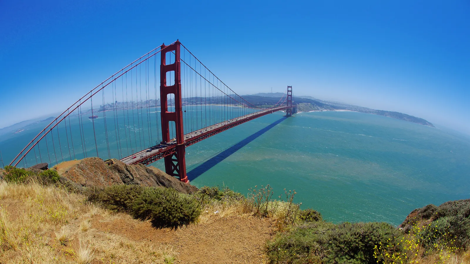 The Golden Gate Bridge spans San Francisco Bay with the city skyline in the distance under a clear blue sky.