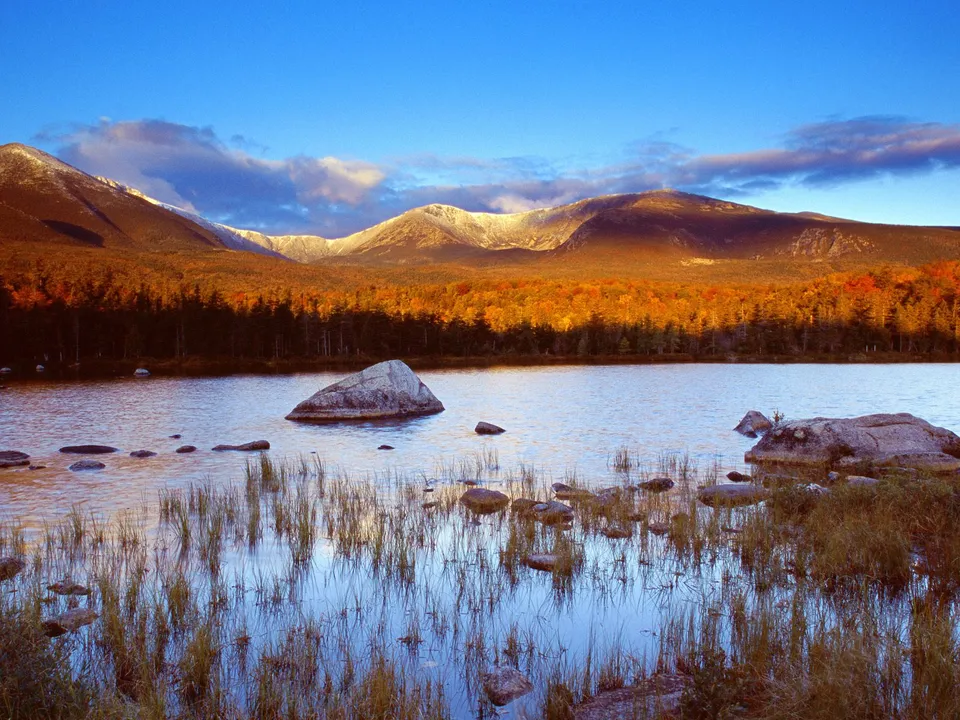 Late afternoon sun illuminates autumn foliage on mountains reflected in a lake.