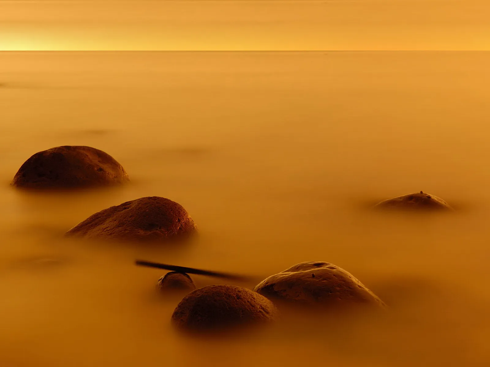 Smooth, rounded rocks emerge from calm, golden-hued water under a soft sky.