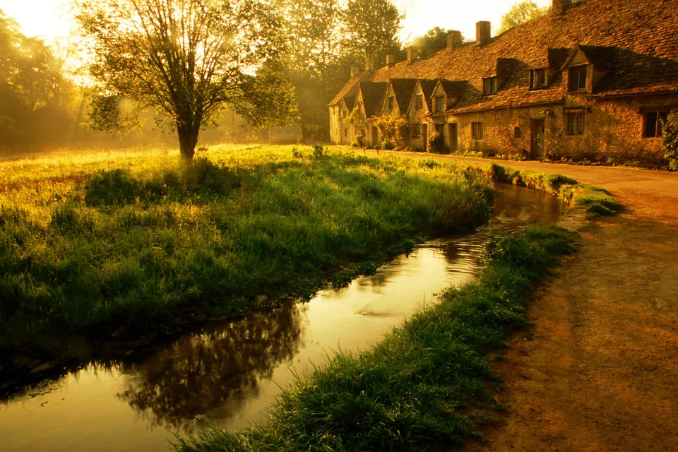 A quaint village with stone cottages and thatched roofs beside a stream bathed in golden morning light.