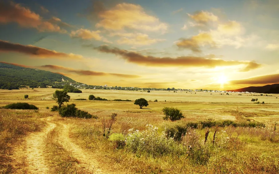 A dirt path winds through dry grass fields towards a distant village under a dramatic sunset sky.