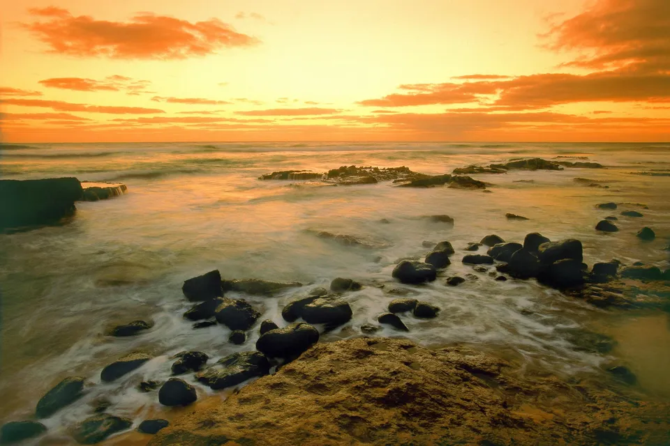 Gentle waves create a misty effect around dark rocks on a rocky shore at sunset.