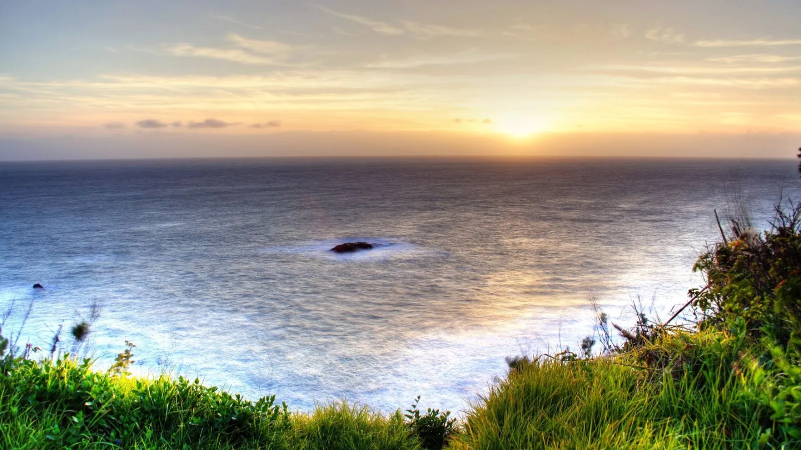 The sun sets over the ocean, casting a golden glow on the water and a distant rock.