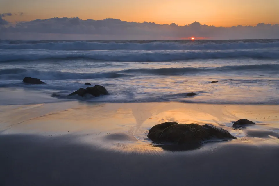The sun sets over the ocean, casting a golden glow on the waves and wet sand.