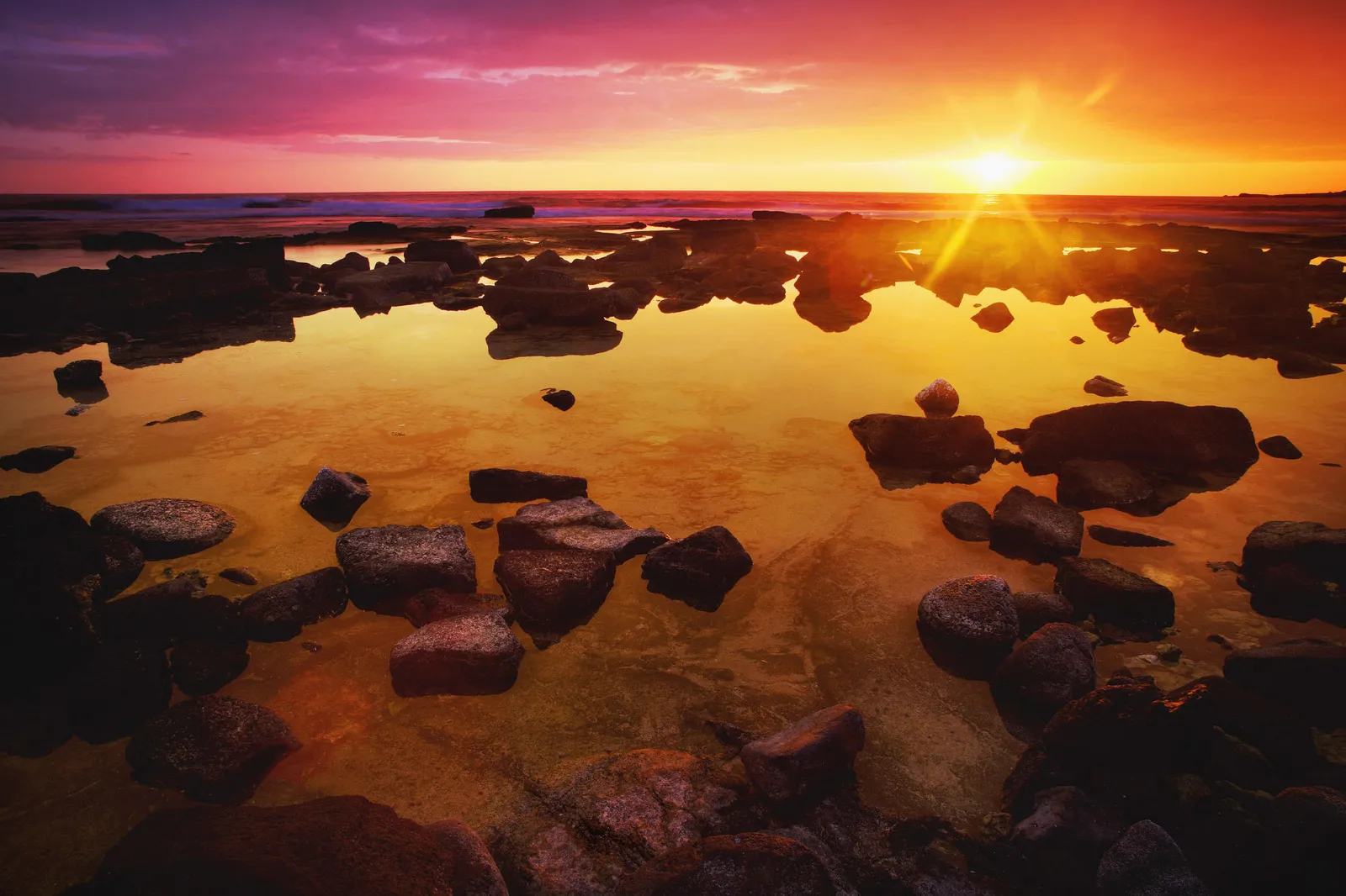 A sunset with pink and yellow hues reflects in tide pools on a rocky shore.