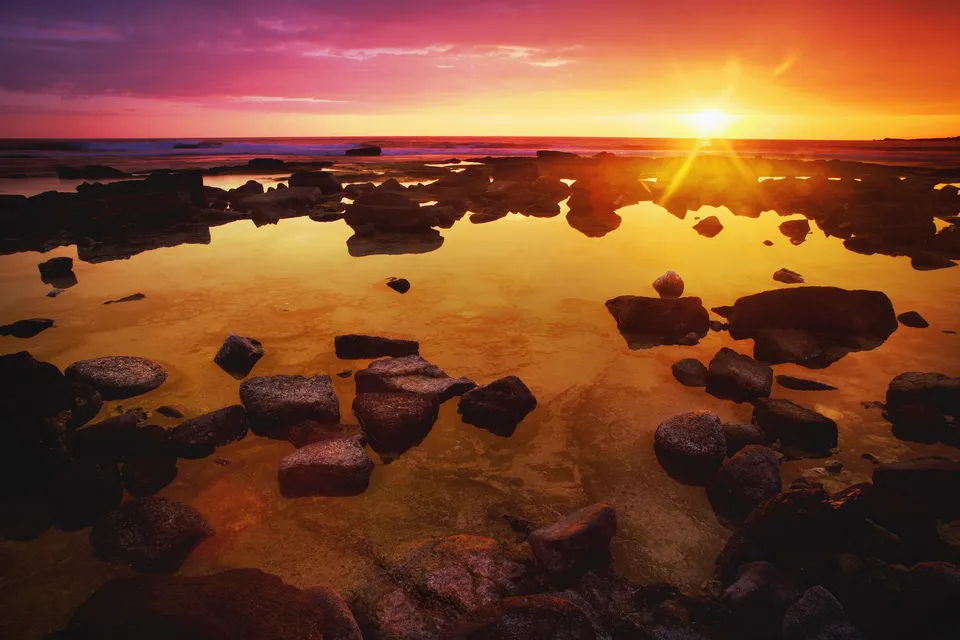 A sunset with pink and yellow hues reflects in tide pools on a rocky shore.
