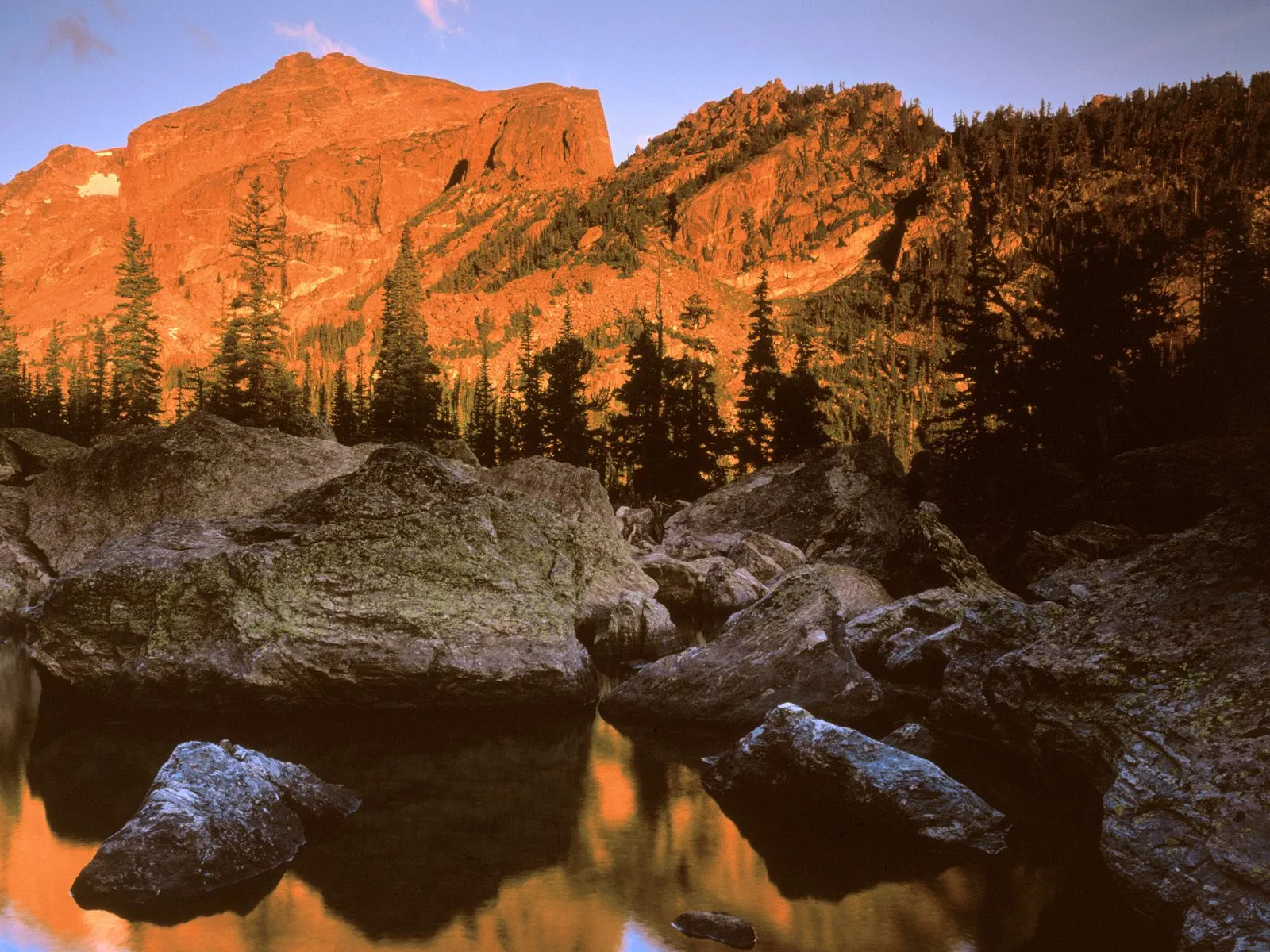 A mountain peak glows orange in the sunrise light, reflected in a calm lake with large rocks in the foreground.