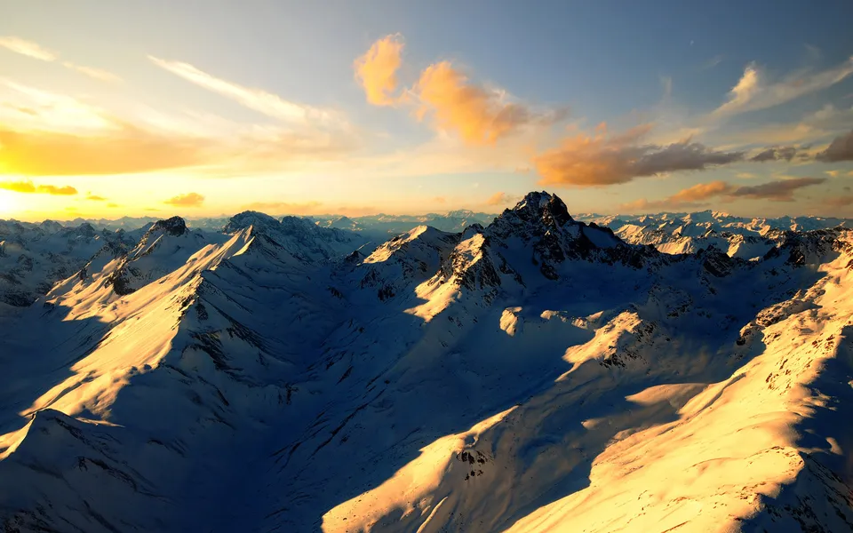 Panoramic view of snow-covered Alps mountains during golden hour with long shadows and a bright sunset sky.