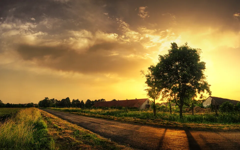 A rural road leads past farm buildings under a dramatic, golden sunset sky.