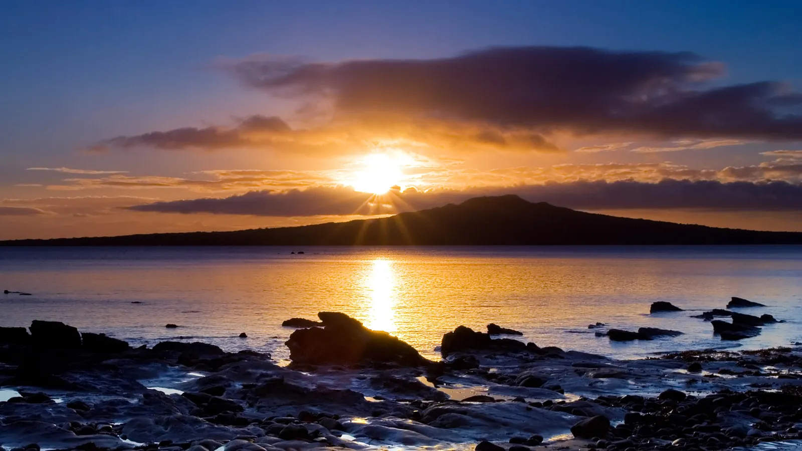 The sun sets behind Rangitoto Island, creating a golden sunburst over the ocean and reflecting on wet rocks in the foreground.