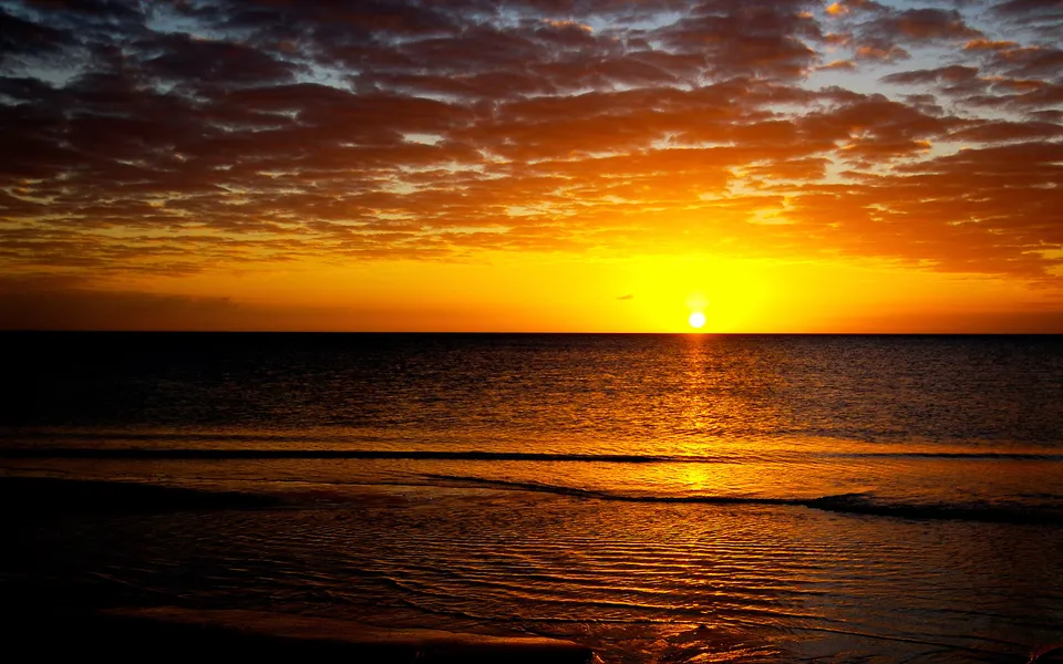 The sun sets over a calm ocean, casting a golden glow on the water and clouds.