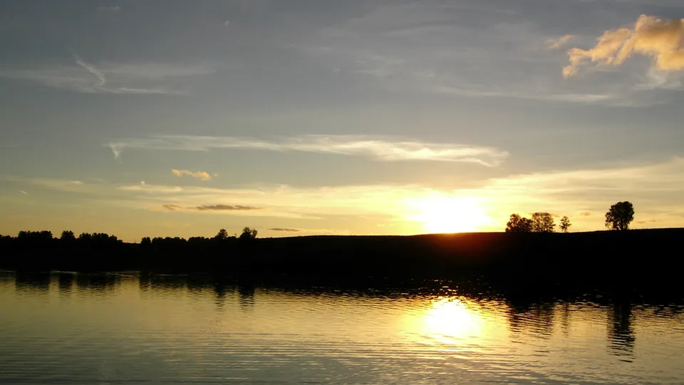 A golden sunset reflects on a calm lake with a silhouetted treeline and wispy clouds.