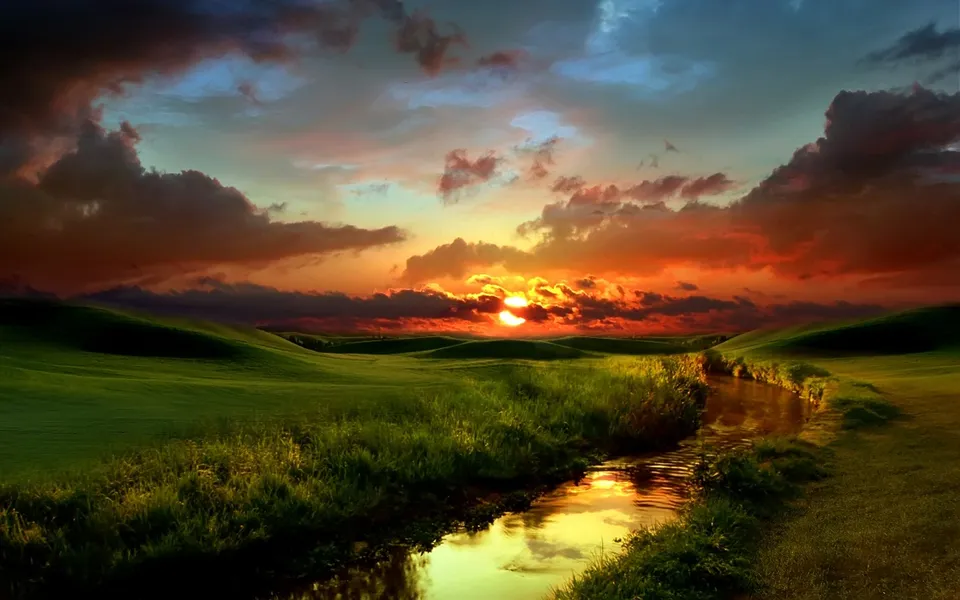 The setting sun casts intense orange and red light, reflected in a stream running through lush green hills.