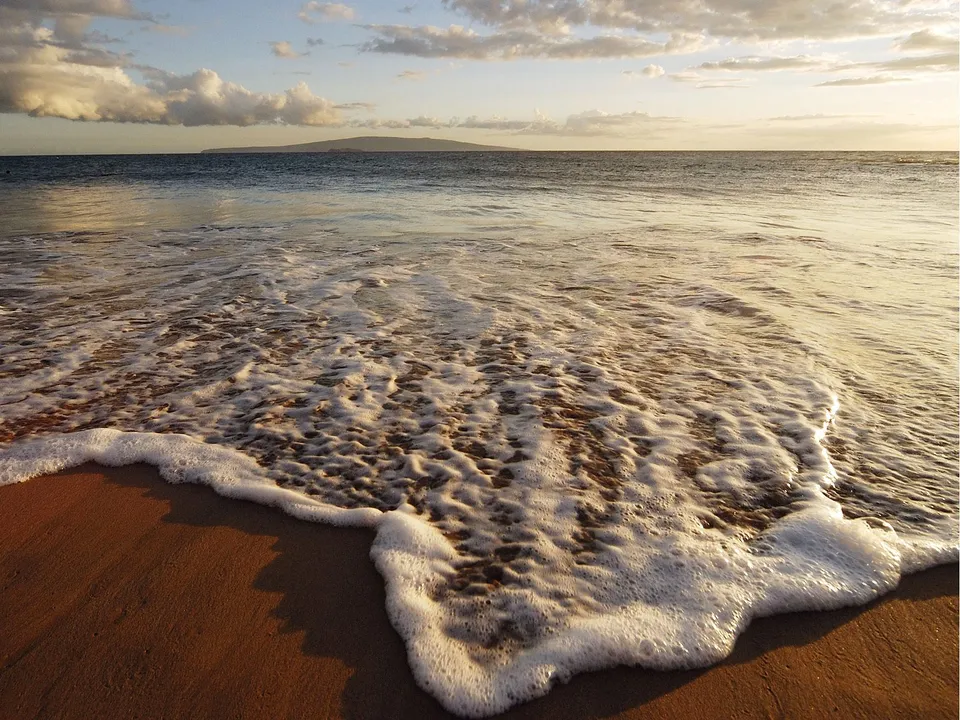 Golden light illuminates gentle waves receding from a sandy shore with a distant island silhouetted against the sky.