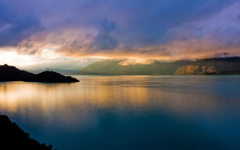 Golden light illuminates distant mountains and clouds over a calm lake at sunset.