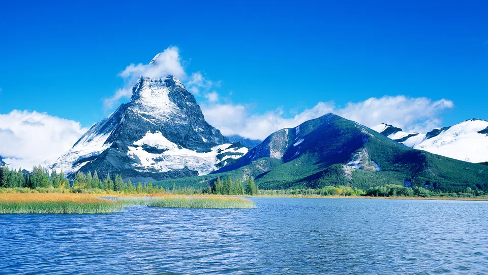 A majestic snowy mountain peak is reflected in a calm lake with golden reeds in the foreground.