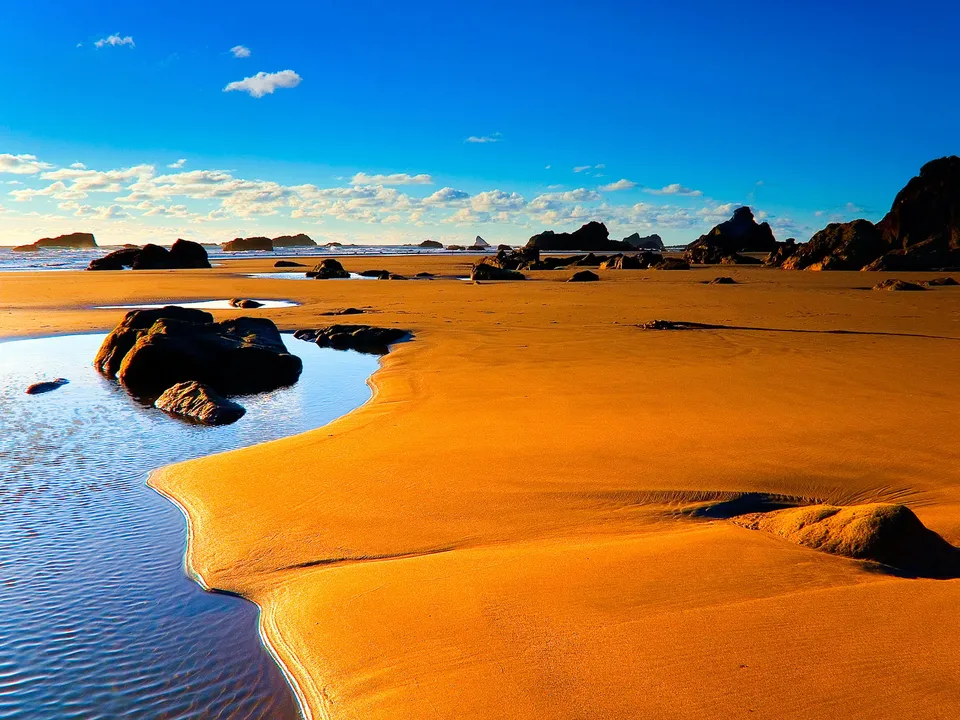 A wide sandy beach with dark rocks and tide pools under a bright blue sky with scattered clouds.