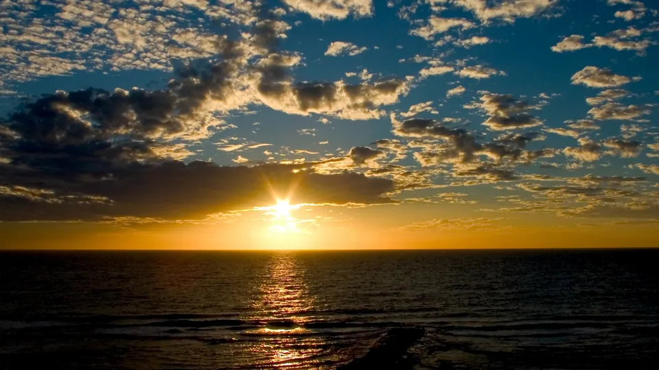 The sun sets on the horizon, casting golden light over the calm ocean and illuminated clouds.