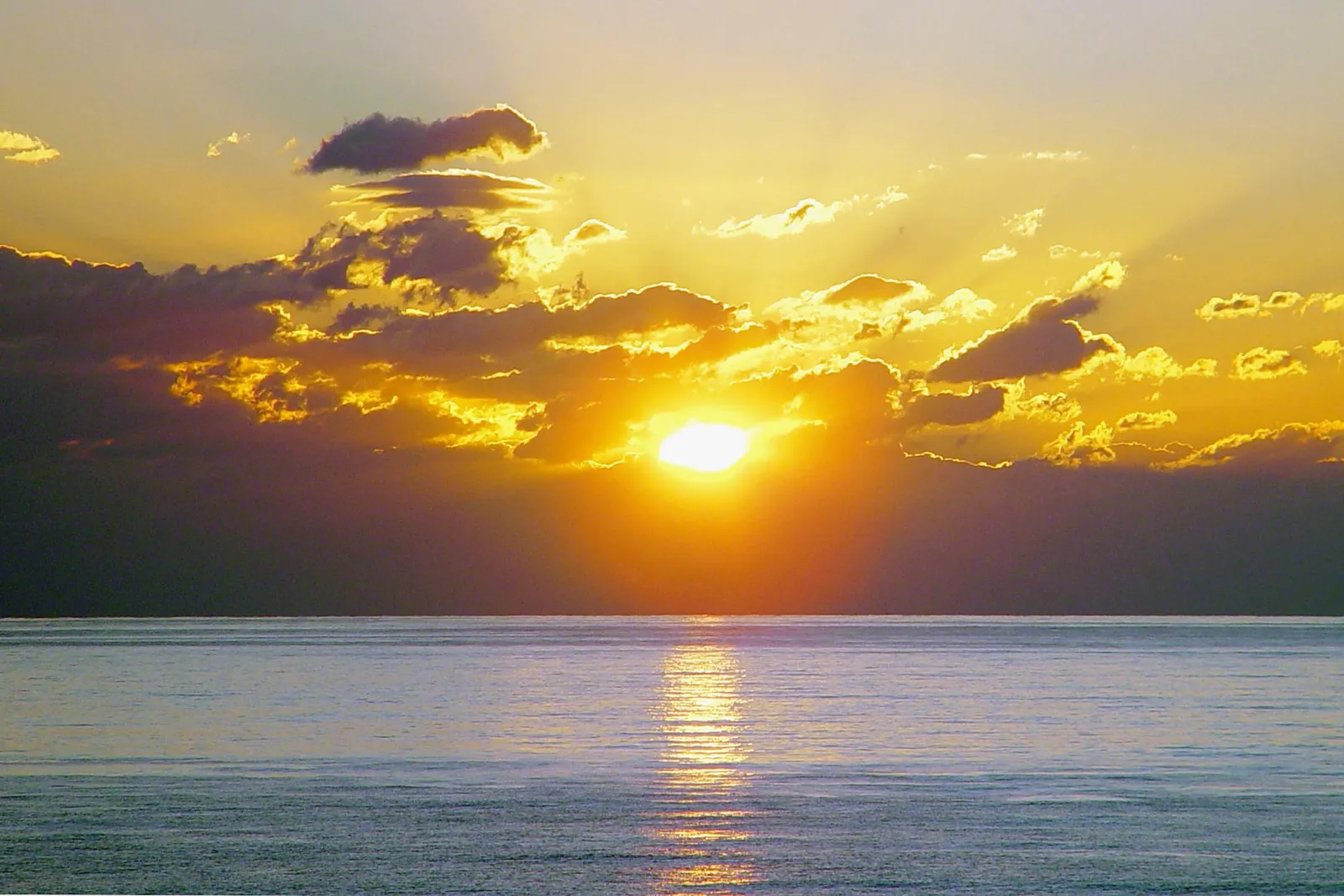 The sun sets over a calm ocean, casting golden rays through dramatic clouds.